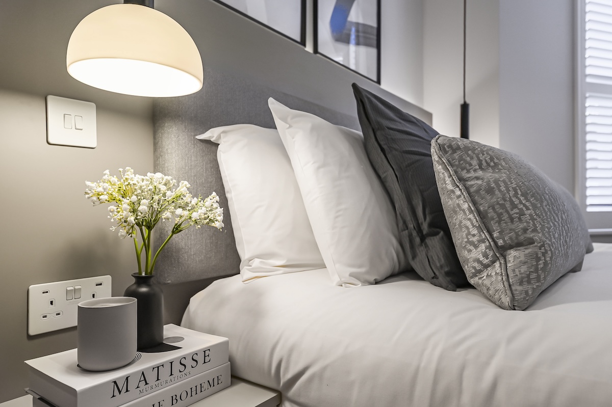 A modern, neatly made bed with white and grey pillows, a bedside table holding stacked books, a vase with white flowers, a grey cup, and a hanging globe lamp illuminating the cosy scene.