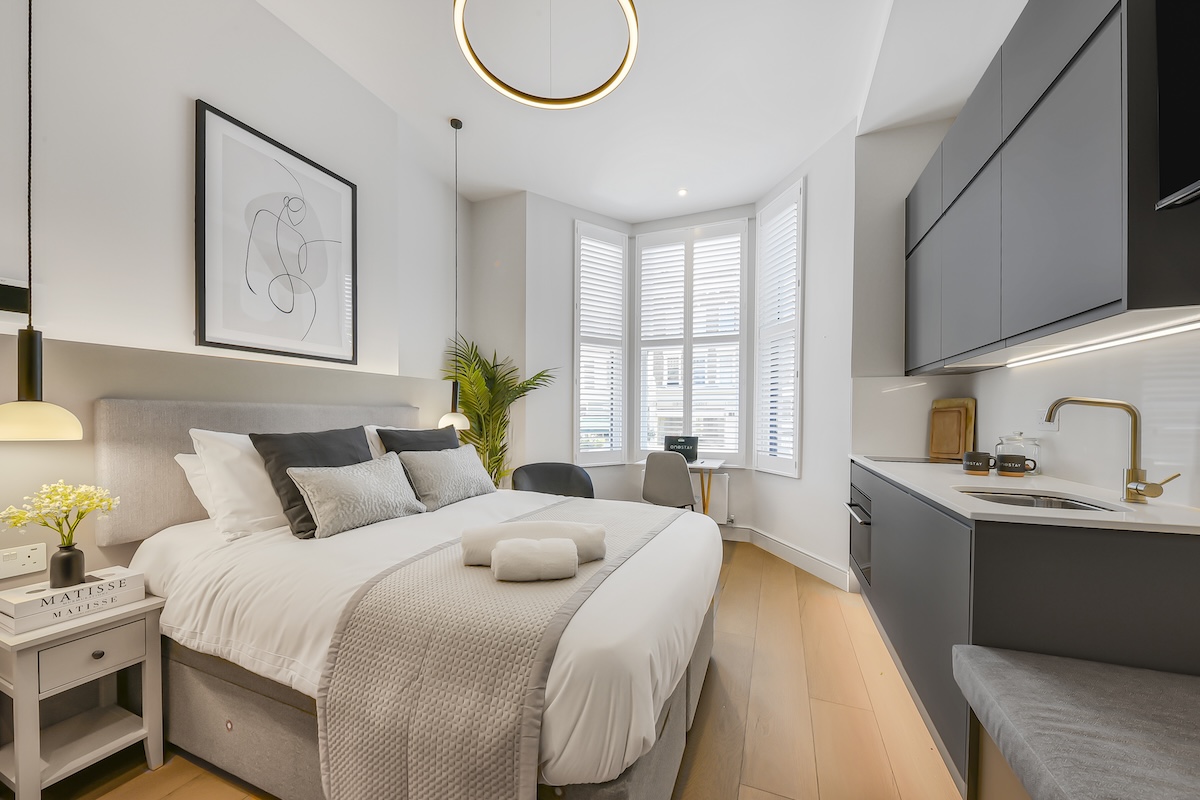 A modern studio flat with a cosy bed, small kitchenette, desk by a bay window with shutters, neutral decor, and soft lighting. Towels are placed on the bed and a plant sits in the corner near the window.