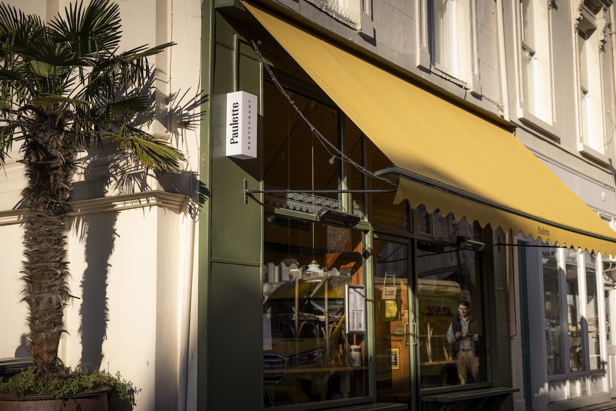 A shopfront with a yellow awning and green trim is pictured in sunlight. A sign reads “Pastifico.” A potted palm tree is on the left, and a person is visible inside through the window.