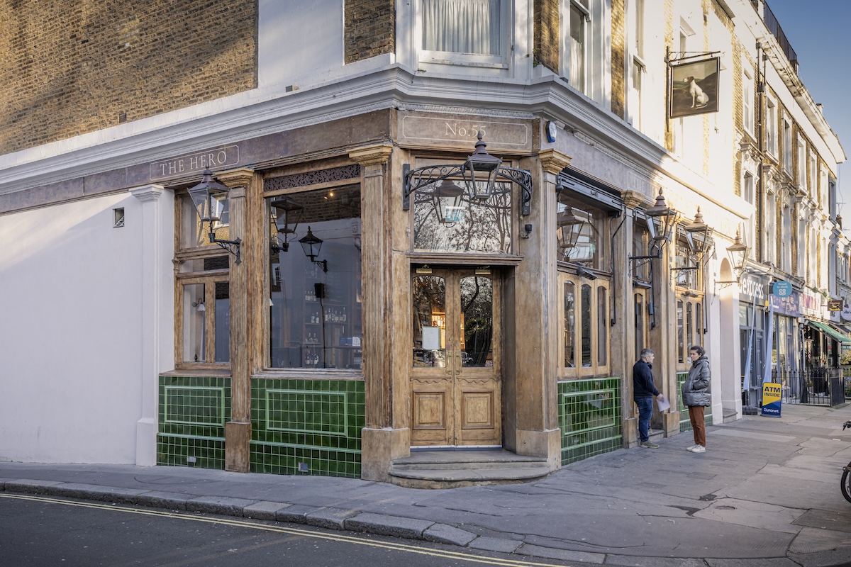 A traditional corner pub with wooden frames, large windows, and green tiled walls. Two people stand outside THE HERO as featured in The One Stay Journal. The quiet street has other shops visible along the pavement.