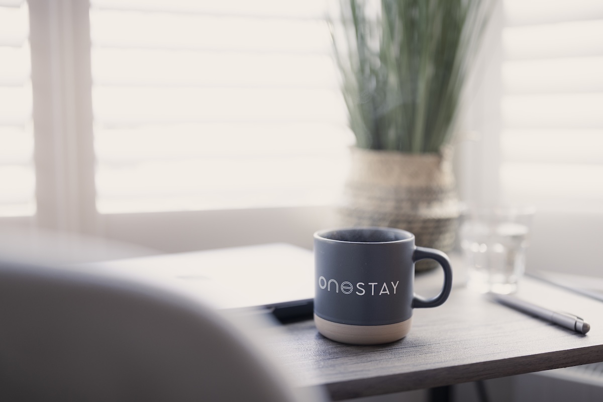 A dark mug labelled oneSTAY sits on a desk next to a pen, a laptop, and a glass of water, with a potted plant and window blinds in the bright background.