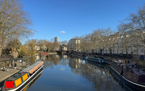 A peaceful canal scene with houseboats moored along both banks, leafless trees, and clear blue sky. People walk on the left path; city buildings are visible in the background.