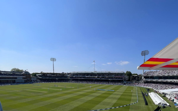 A wide view of a cricket stadium on a sunny day, with a match in progress. The stands are full of spectators, and the sky is clear and blue.