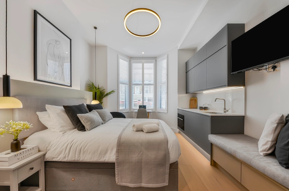 Modern studio flat with a neatly made bed, grey cupboards, a wall-mounted TV, a kitchenette, a window with white shutters, cosy lighting, and minimalistic decor.