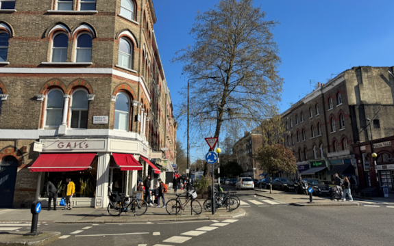 A sunny street corner with people walking, bicycles parked, and shops including GAIL'S bakery. Tall trees and brick buildings line both sides of the street under a clear blue sky.