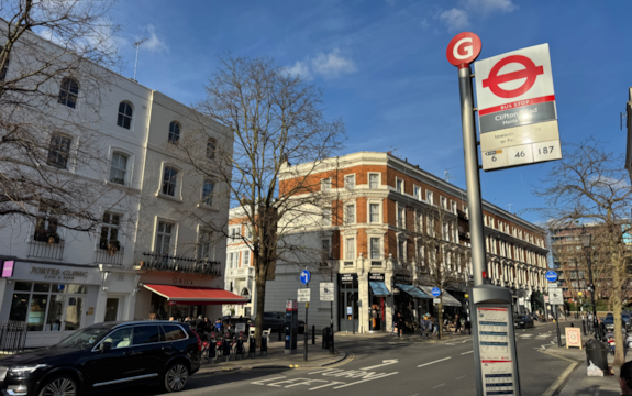 A street scene shows a London bus stop sign with route numbers, shops, parked cars, and people sitting outside a café under a blue sky with some trees and white buildings in the background.